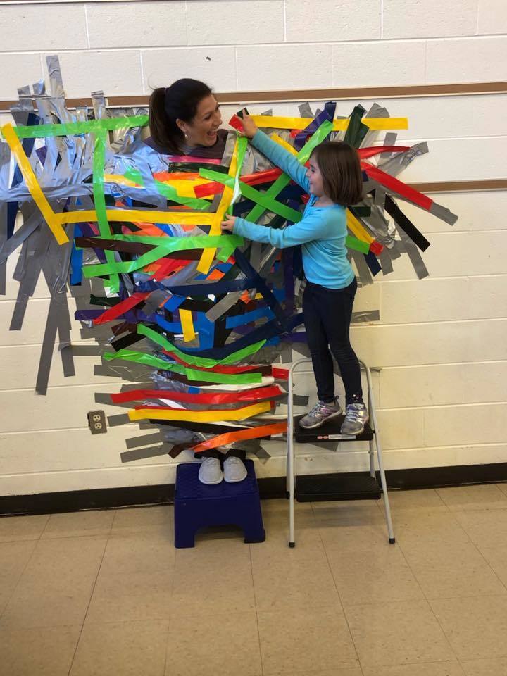 Ms. Kelly gets taped to a wall after a fundraiser. Please note: photos were taken prior to pandemic and masking mandates.