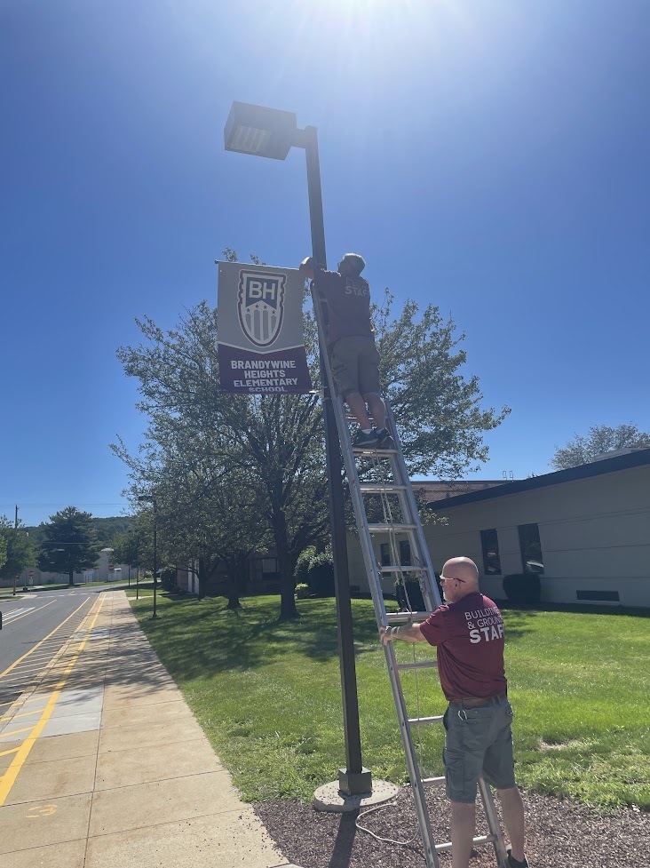 Two maintenance workers hang an avenue banner on a lamp post that says 'Brandywine Heights Elementary School'